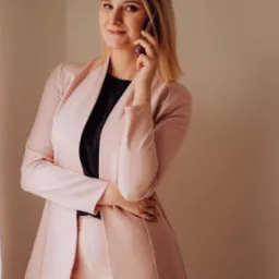 Uśmiechnięta blondynka w różowym garniturze rozmawia przez telefon, opierając rękę na biodrze, na neutralnym tle.