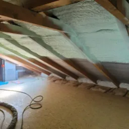 Poddasze z drewnianą konstrukcją dachu po natrysku izolacji, widoczne przewody i niebieska mata ochronna na podłodze.