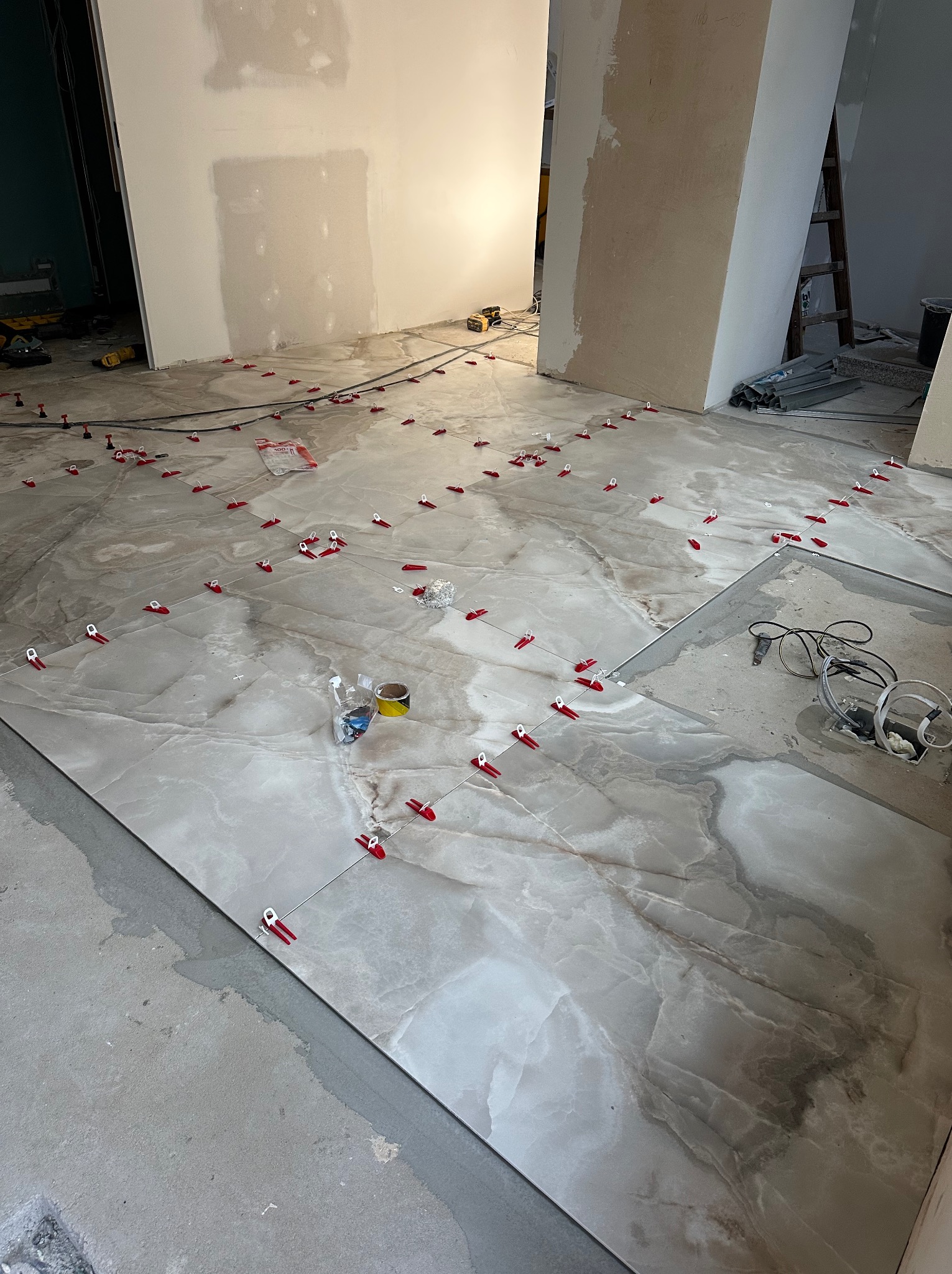 Układanie płytek podłogowych z użyciem systemu poziomowania. Widoczne płytki imitujące marmur, czerwone klipsy i kliny. W tle surowe ściany w trakcie wykończenia.