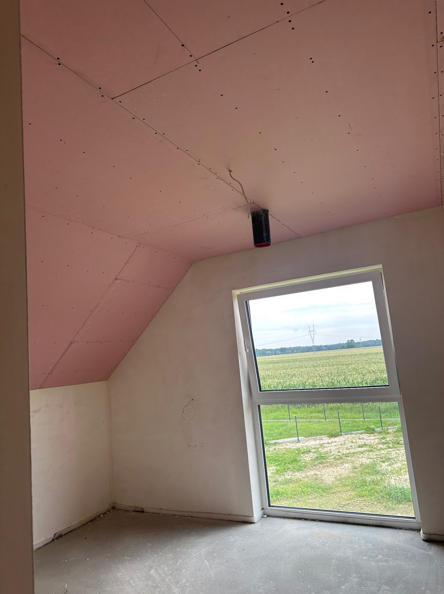 Poddasze z oknem po montażu płyt gipsowo-kartonowych na skosach dachu. Widoczne łączenia płyt i wystający przewód wentylacyjny. Za oknem zielony krajobraz.