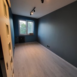 Patryk Mikulski - Pomalowane na ciemny odcień szarości ściany w pustym pokoju z panelami podłogowymi, widoczne czarne reflektory sufitowe i okno z grzejnikiem.