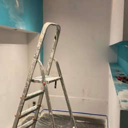 Patryk Mikulski - Pokój w trakcie odświeżania: ściany częściowo pomalowane na biało, jedna ściana z turkusowym akcentem i abstrakcyjnym wzorem, podłoga zabezpieczona folią malarską, drabina malarska w centrum kadru.