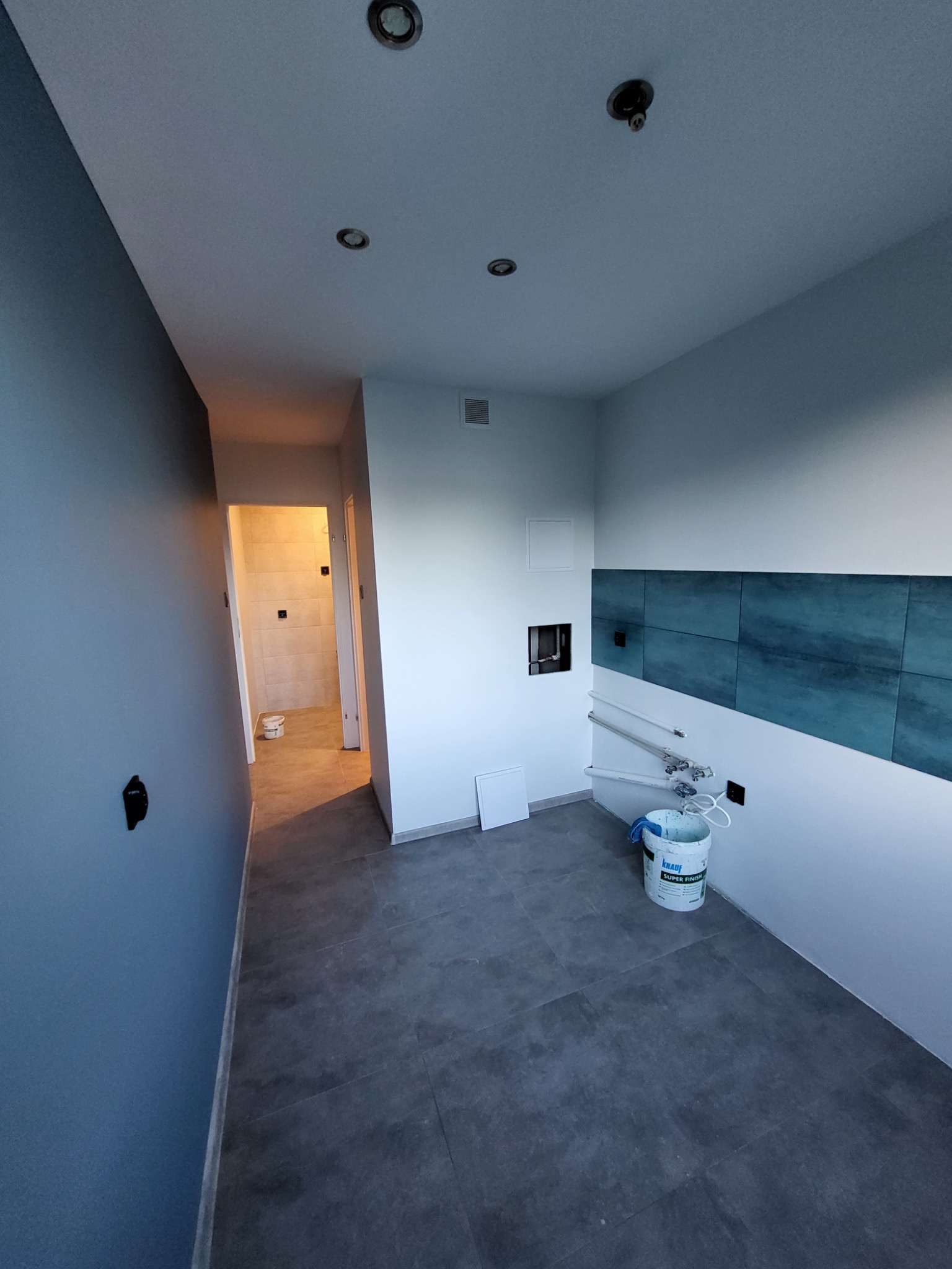 Wnętrze w trakcie prac wykończeniowych: pomalowana na niebiesko ściana, fragment ściany z glazurą w odcieniach błękitu, widoczne puszki z materiałami budowlanymi oraz fragment wykończonej łazienki...