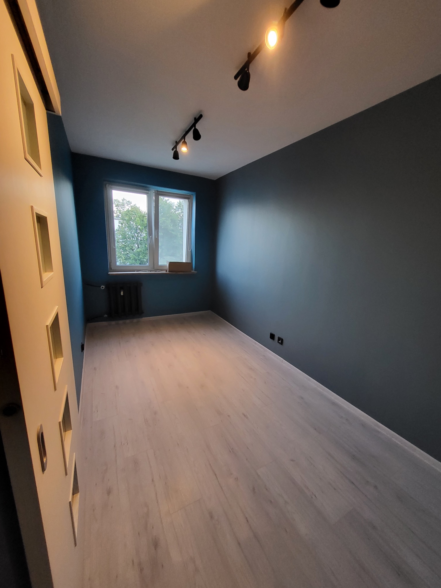 Pomalowane na ciemny odcień szarości ściany w pustym pokoju z panelami podłogowymi, widoczne czarne reflektory sufitowe i okno z grzejnikiem.