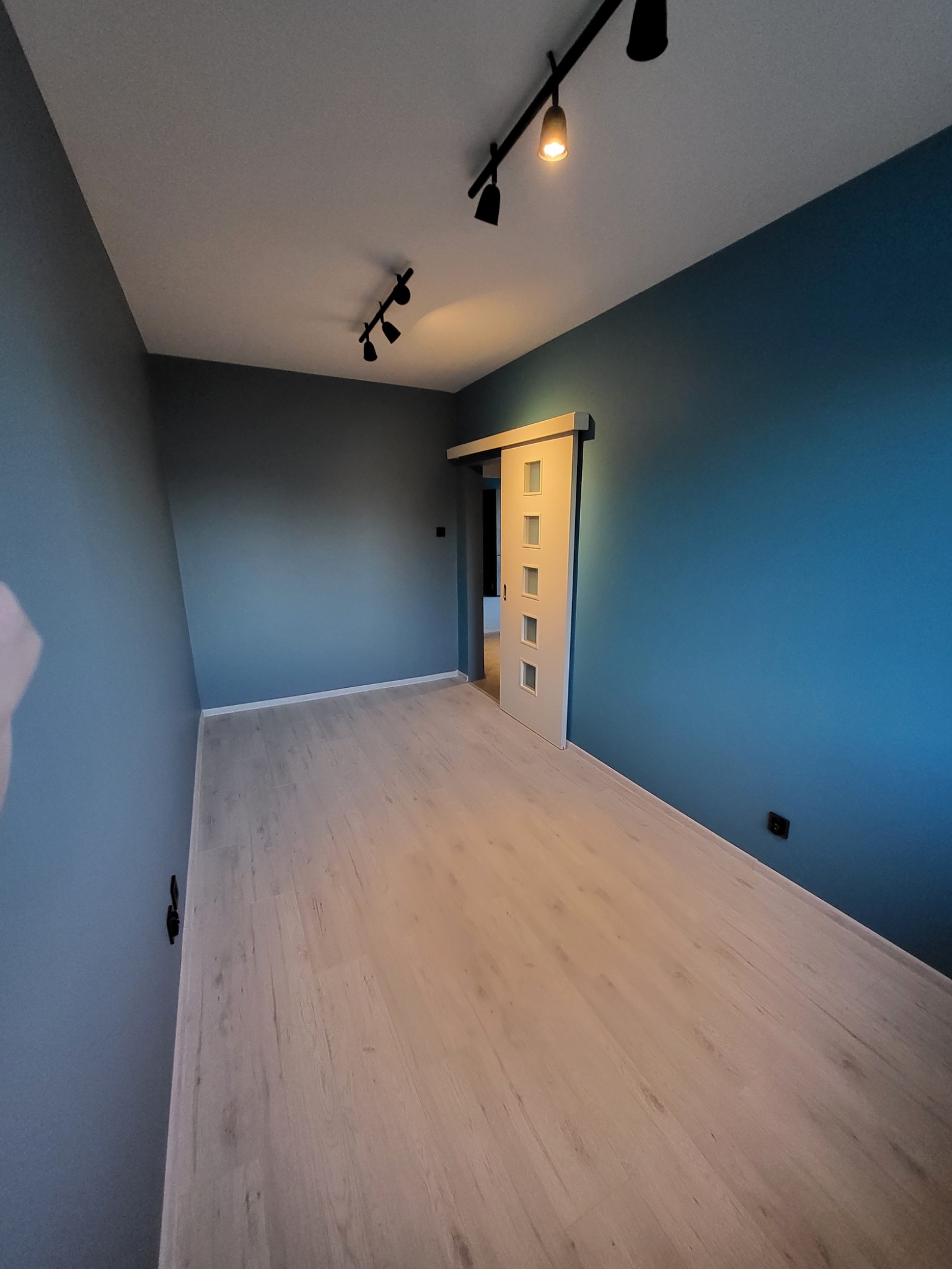 Pomalowane na dwa odcienie szarości i błękitu ściany, jasne panele podłogowe oraz białe, przesuwne drzwi z szybkami w wyremontowanym pokoju w Aleksandrowie Łódzkim.