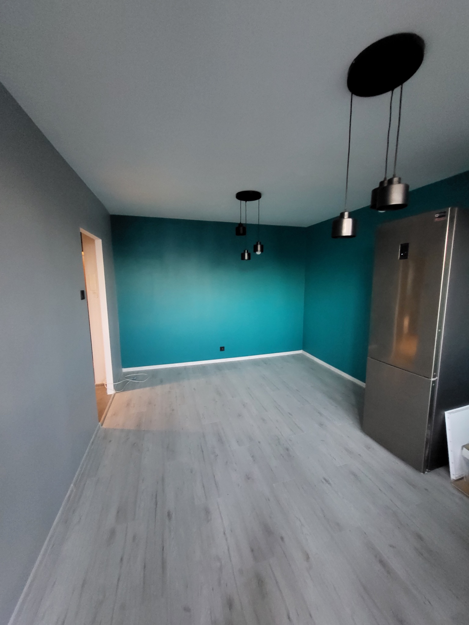 Wnętrze po remoncie: szara ściana kontrastuje z turkusową, nowoczesne lampy wiszące, panele podłogowe imitujące drewno, widoczna lodówka ze stali nierdzewnej.