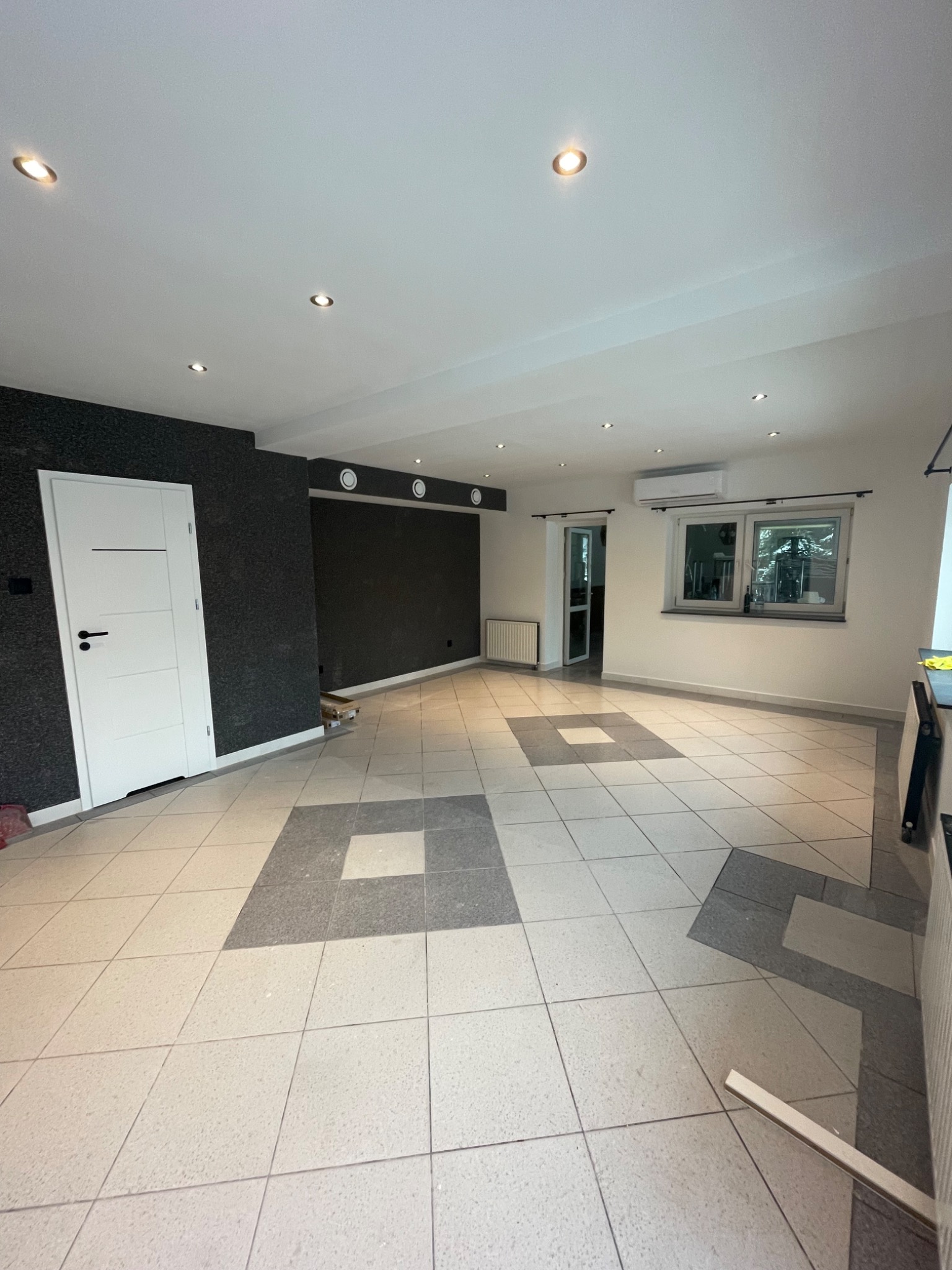 Przestronne wnętrze po remoncie z jasnymi płytkami podłogowymi w szary kwadratowy wzór, ciemną ścianą z fakturą i białymi drzwiami oraz oświetleniem punktowym w suficie.