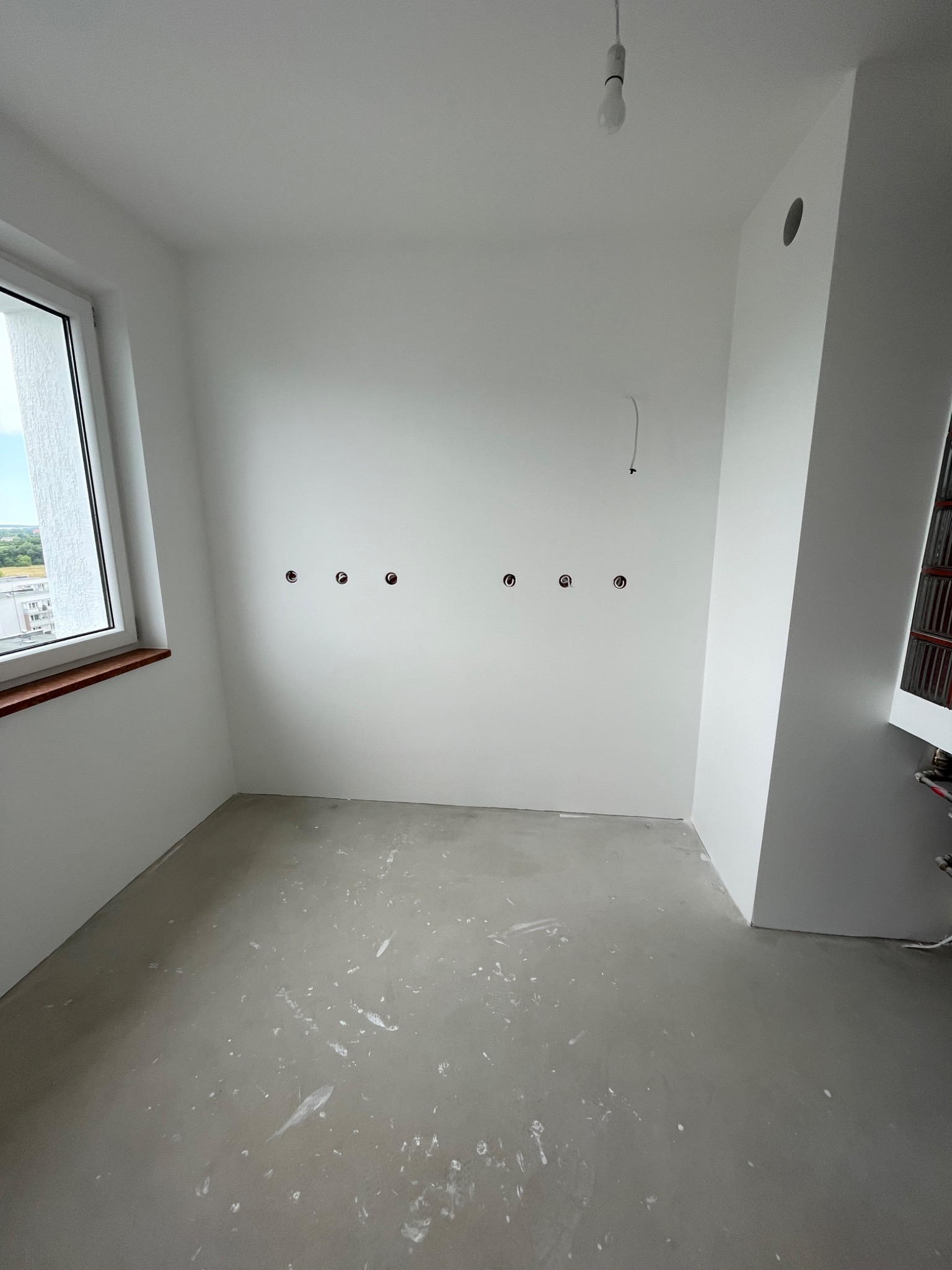 Pusty pokój w trakcie remontu, świeżo wygładzone ściany pomalowane na biało, surowa wylewka na podłodze, widoczne puszki elektryczne w ścianie i wisząca żarówka.