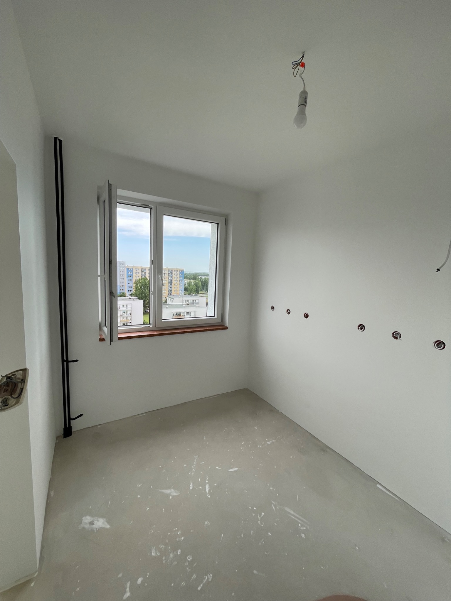 Pusty pokój w stanie deweloperskim z otwartym oknem, widok na osiedle, ślady po pracach budowlanych na podłodze, wystające kable z sufitu, przygotowane gniazdka elektryczne.
