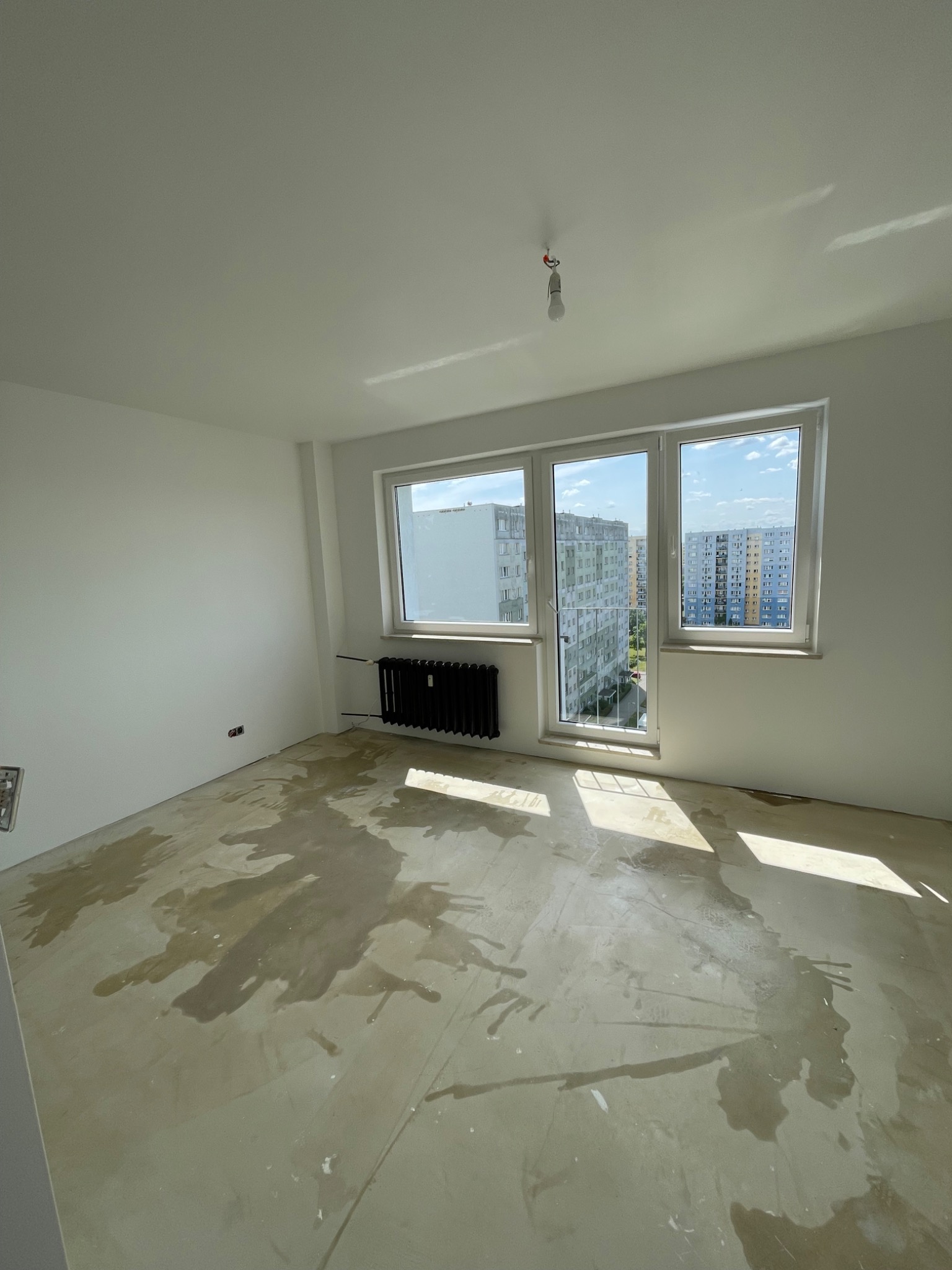 Pokój w trakcie prac przygotowawczych do wykończenia, z widocznymi śladami wilgoci na podłodze, świeżo pomalowanymi ścianami na biało i grzejnikiem pod oknem.