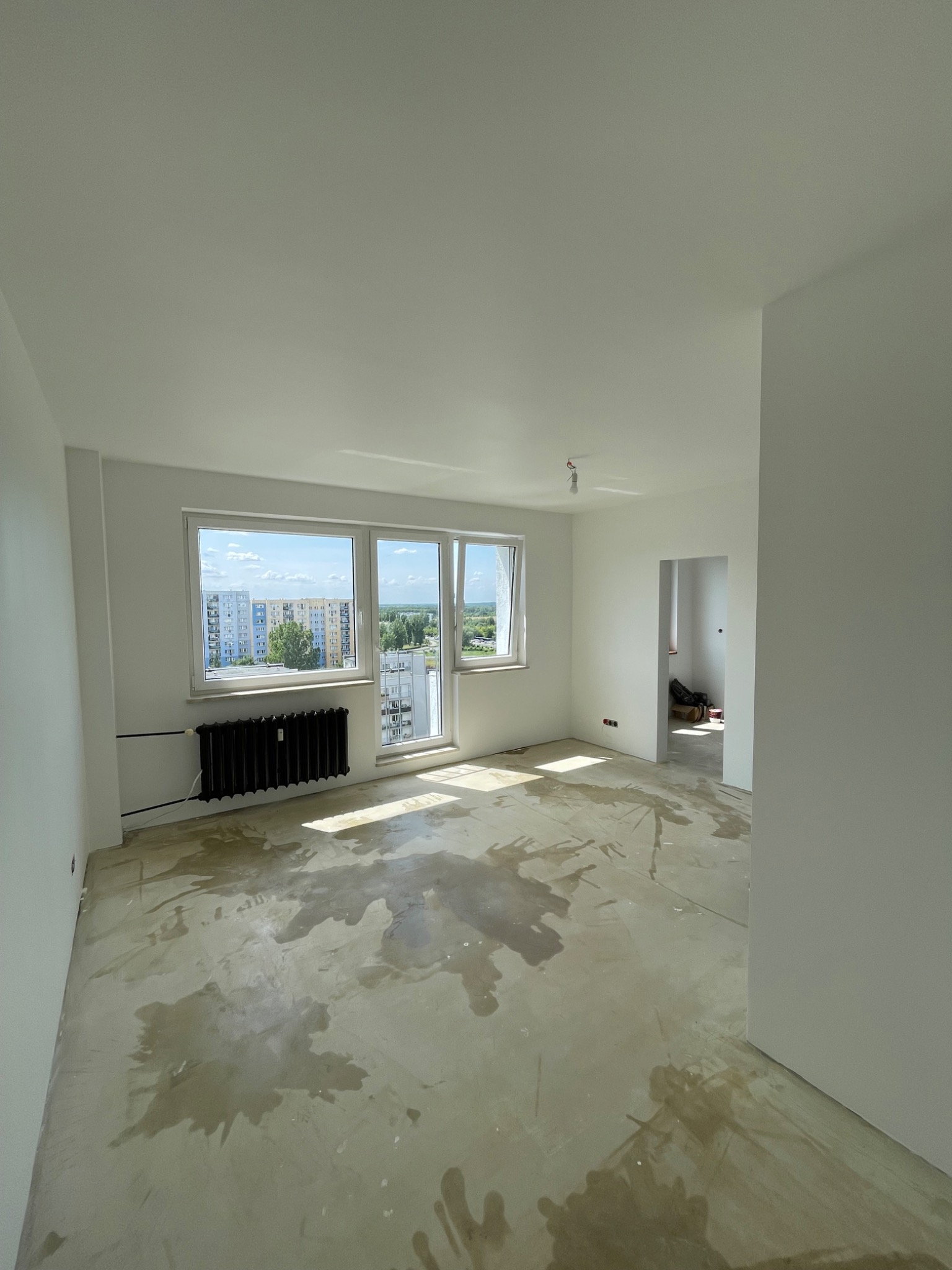 Wnętrze pustego, odświeżonego pokoju z białymi ścianami, z widocznymi śladami wilgoci na podłodze przed dużym oknem z widokiem na blokowisko w oddali. Czarny, żeliwny grzejnik zamontowany pod oknem...