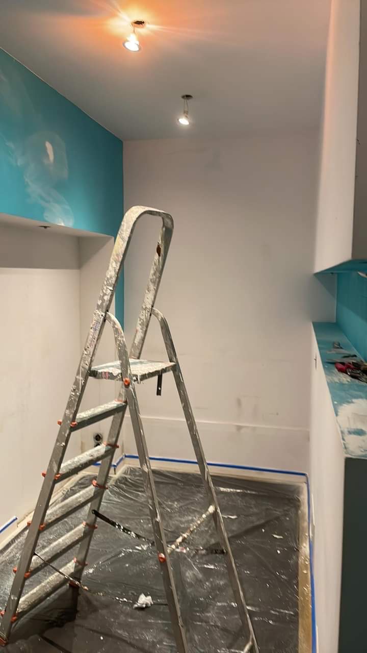 Pokój w trakcie odświeżania: ściany częściowo pomalowane na biało, jedna ściana z turkusowym akcentem i abstrakcyjnym wzorem, podłoga zabezpieczona folią malarską, drabina malarska w centrum kadru.