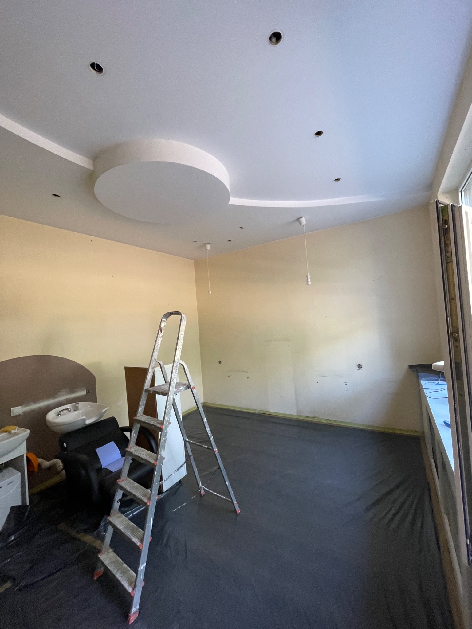 Pokój w trakcie prac przygotowawczych: zabezpieczona folią podłoga, drabina aluminiowa, surowe ściany w odcieniach beżu z widocznymi otworami po oświetleniu, dekoracyjny okrąg na suficie.