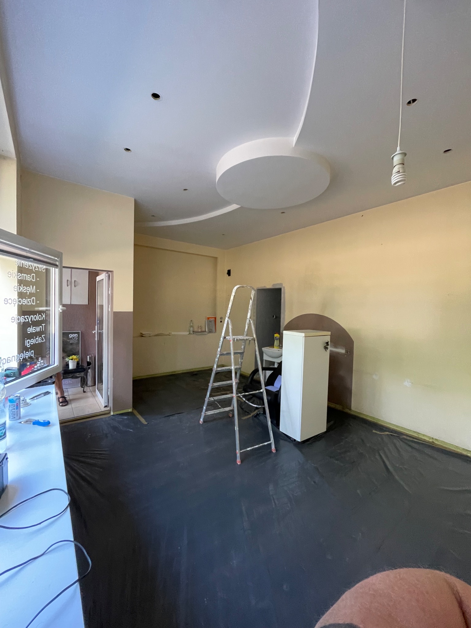 Przygotowane pomieszczenie do prac wykończeniowych: podłoga zabezpieczona czarną folią, ściany w odcieniach beżu, widoczna drabina aluminiowa, dekoracyjny okrągły element na suficie i wisząca...