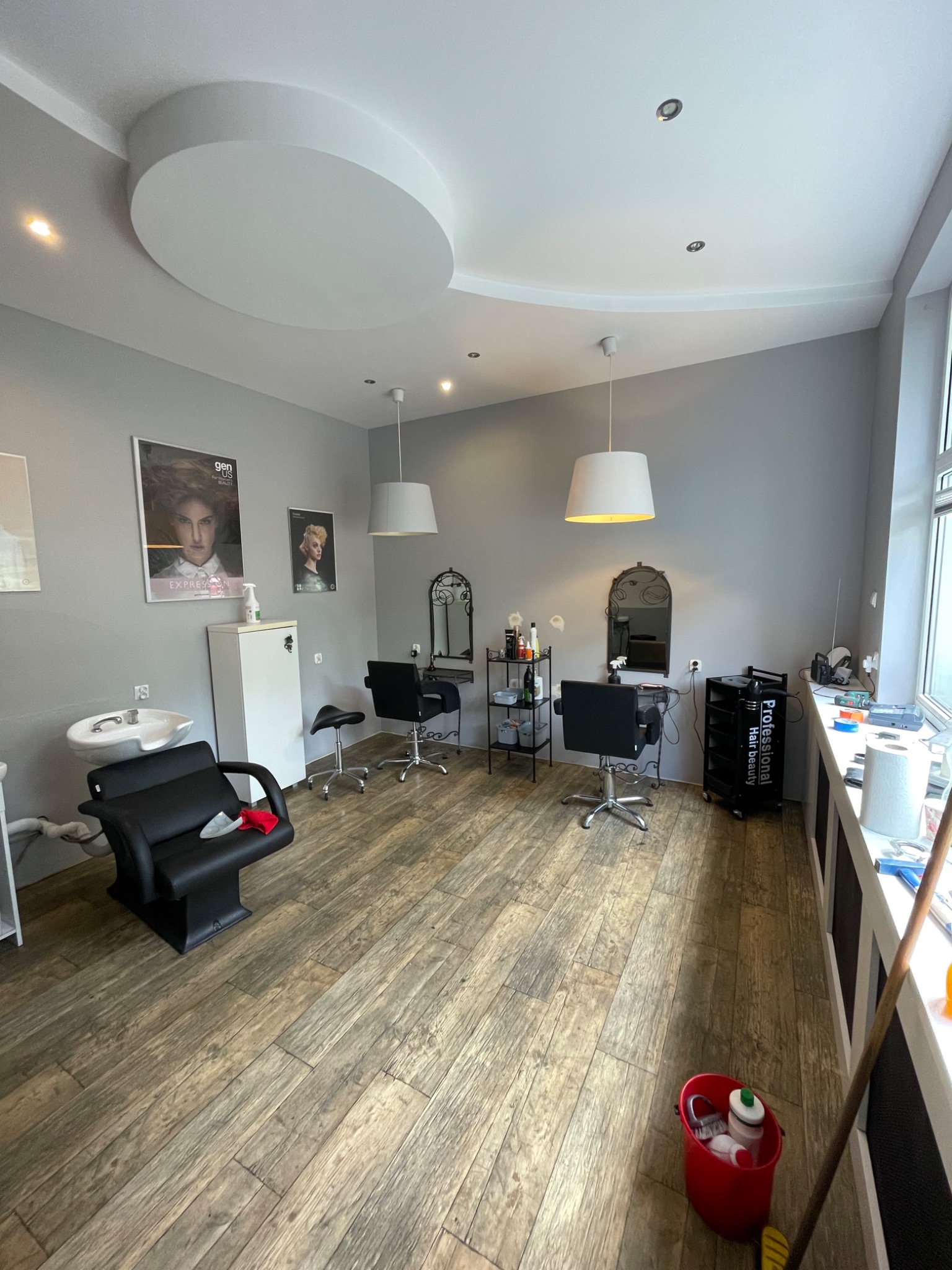 Jasne wnętrze salonu fryzjerskiego z drewnopodobną podłogą, wyposażone w stanowiska z lustrami, czarne fotele fryzjerskie, umywalkę i wiszące lampy z okrągłymi kloszami, dekorowane plakatami...