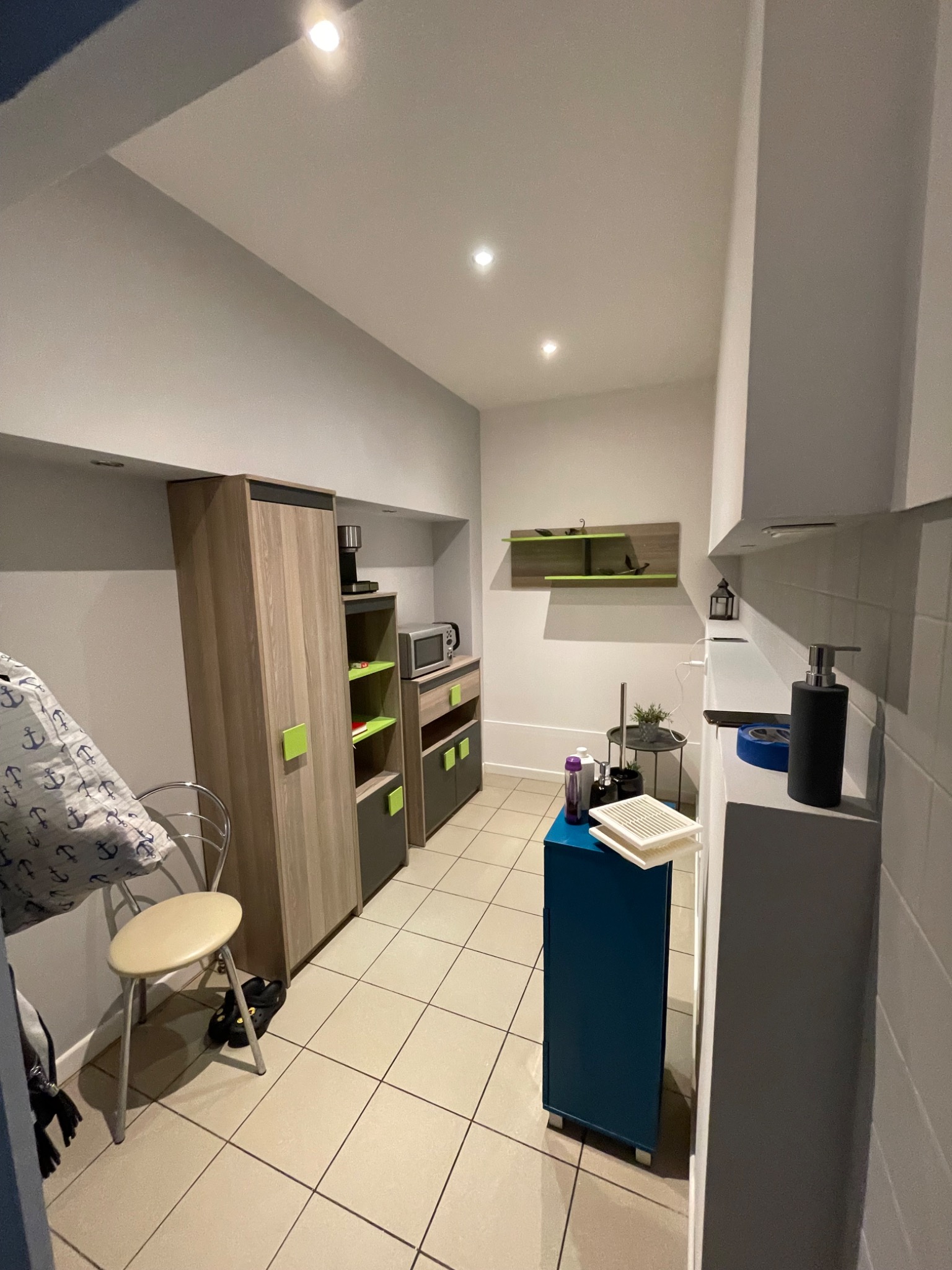 Wnętrze małej kuchni po remoncie, z nowoczesnymi meblami w odcieniach szarości i drewna, jasnymi ścianami i ceramiczną podłogą w neutralnych barwach. Na pierwszym planie niebieska szafka...