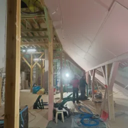 Montaż płyt na poddaszu: Dwóch pracowników w trakcie prac wykończeniowych, widoczne drewniane belki konstrukcyjne, niebieskie kable i narzędzia na podłodze, oświetlenie robocze.