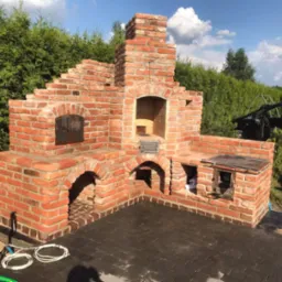 Murowany piec zewnętrzny z cegły klinkierowej, z paleniskiem i wędzarnią, wykończony na zewnątrz, z widoczną instalacją wodną i elektryczną oraz fragmentem samochodu w tle.