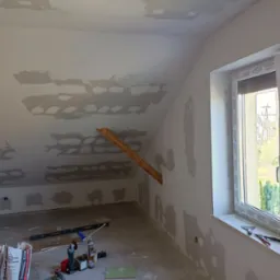 Poddasze w trakcie prac wykończeniowych: ściany i sufit po szpachlowaniu, widoczne ślady po łatach, narzędzia i materiały budowlane rozłożone na podłodze, okno z widokiem na zewnątrz.