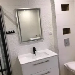 Wykończona łazienka z białą armaturą, umywalką z szafką, podświetlanym lustrem i dekoracyjnymi, trójwymiarowymi płytkami ściennymi w kolorze białym.