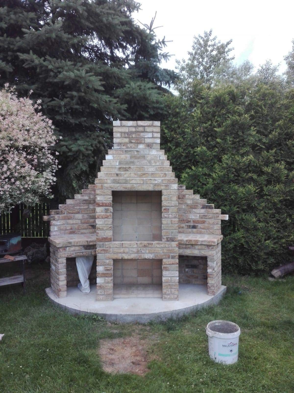 Murowany grill ogrodowy z jasnej cegły, z kominem o schodkowej konstrukcji, na betonowej podstawie, otoczony zielenią; widoczny pojemnik z resztkami zaprawy.