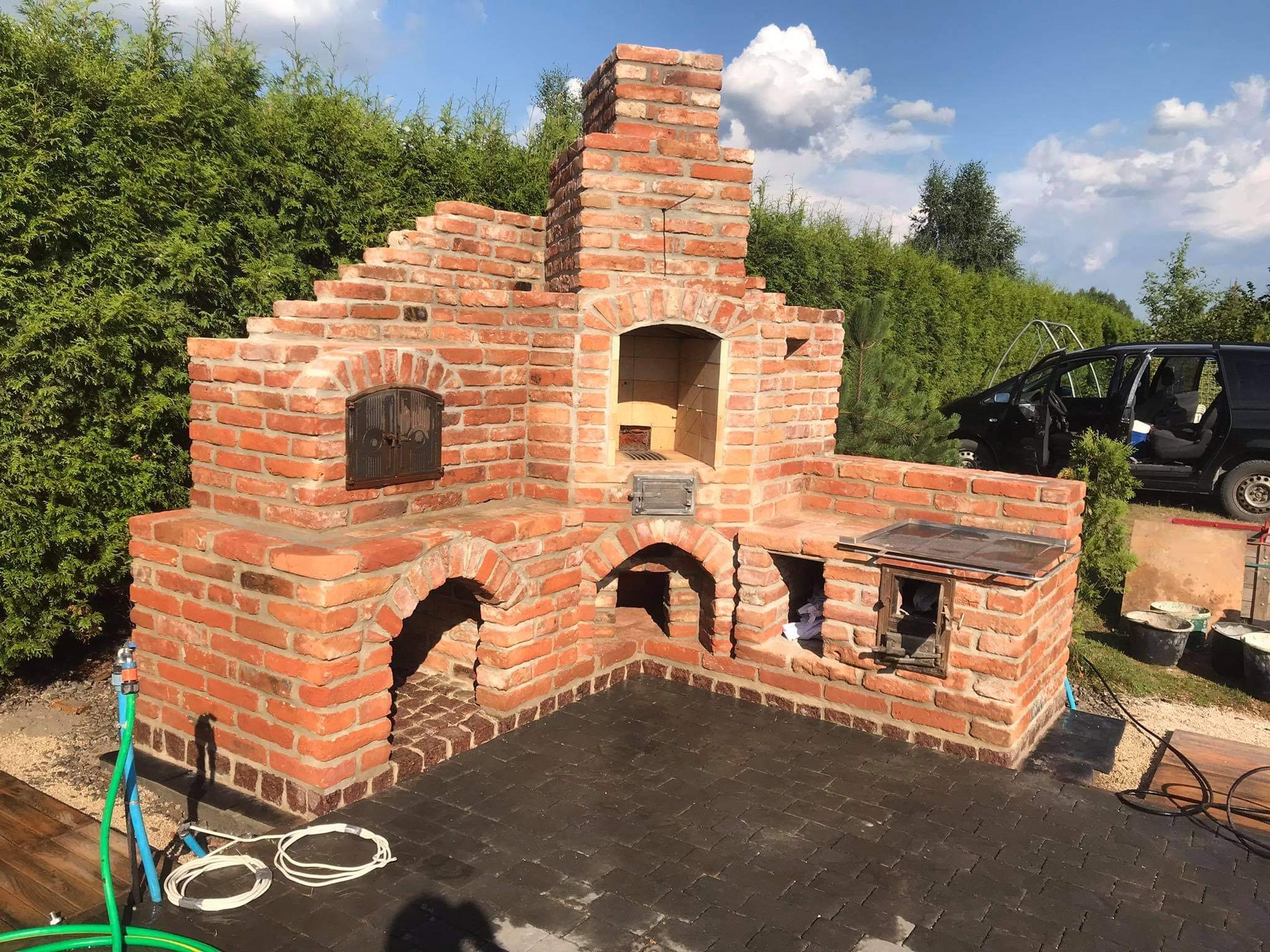 Murowany piec zewnętrzny z cegły klinkierowej, z paleniskiem i wędzarnią, wykończony na zewnątrz, z widoczną instalacją wodną i elektryczną oraz fragmentem samochodu w tle.
