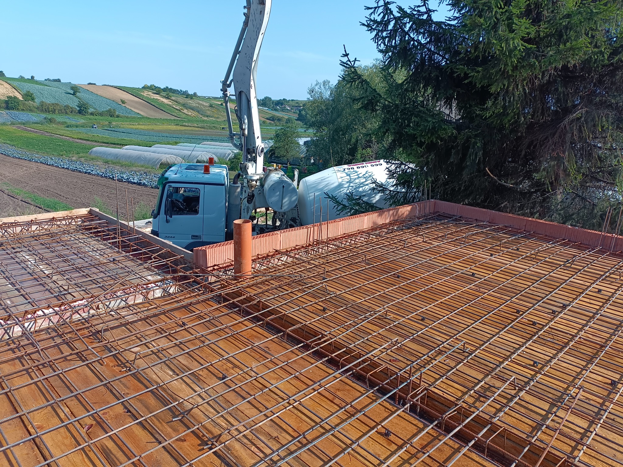 Wylewanie betonu na zbrojenie stropu budynku z użyciem pompy do betonu, na tle widoczne pola uprawne i szklarnie.