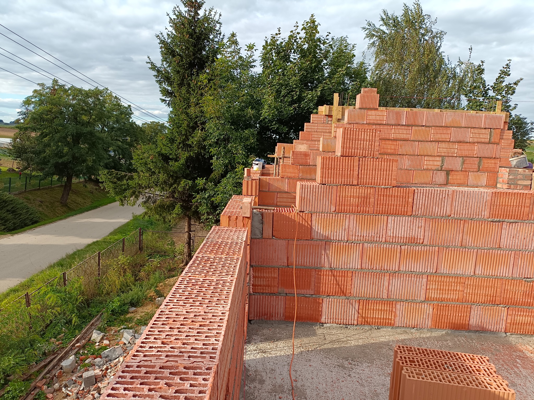 Wznoszenie muru z czerwonej ceramiki poryzowanej na budowie domu jednorodzinnego, widok na okolicę z zielenią i pochmurnym niebem.