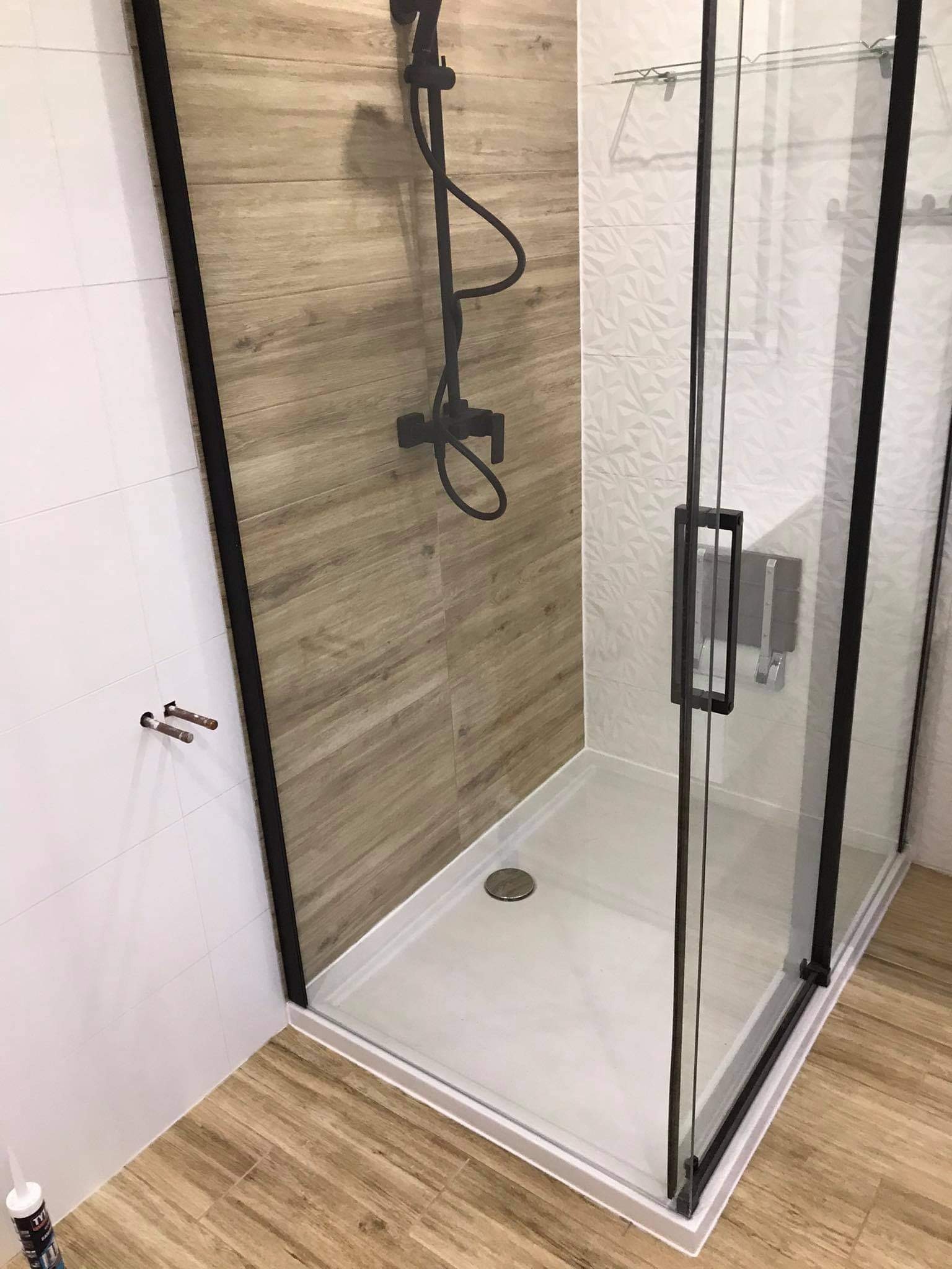 Nowoczesna kabina prysznicowa z czarnymi profilami i deszczownicą, wykończona płytkami imitującymi drewno i białymi płytkami z geometrycznym wzorem. Widoczna butelka silikonu na podłodze.