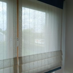 WBW Studio Dekoracyjne Okien - Beżowa, lniana roleta w stylu rzymskim z granatowym wykończeniem, zamontowana w oknie z widokiem na zieleń.