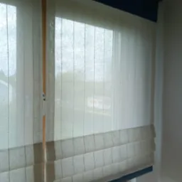 Beżowa, lniana roleta w stylu rzymskim z granatowym wykończeniem, zamontowana w oknie z widokiem na zieleń.