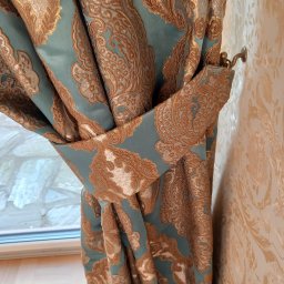 WBW Studio Dekoracyjne Okien - Turkusowa zasłona z ornamentem w kolorze złota, podpięta ozdobnym troczkiem, widoczna na tle ściany w kwiatowe wzory i fragment okna.