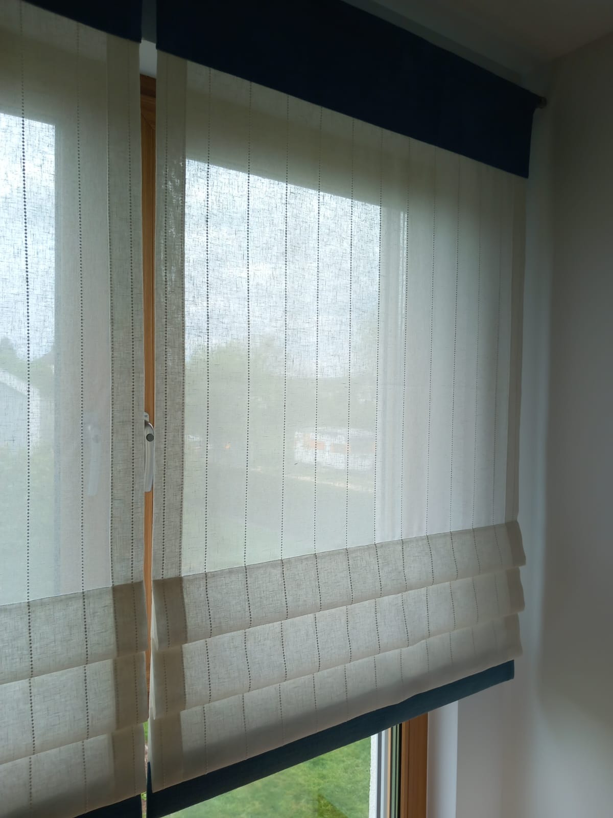 Beżowa, lniana roleta w stylu rzymskim z granatowym wykończeniem, zamontowana w oknie z widokiem na zieleń.