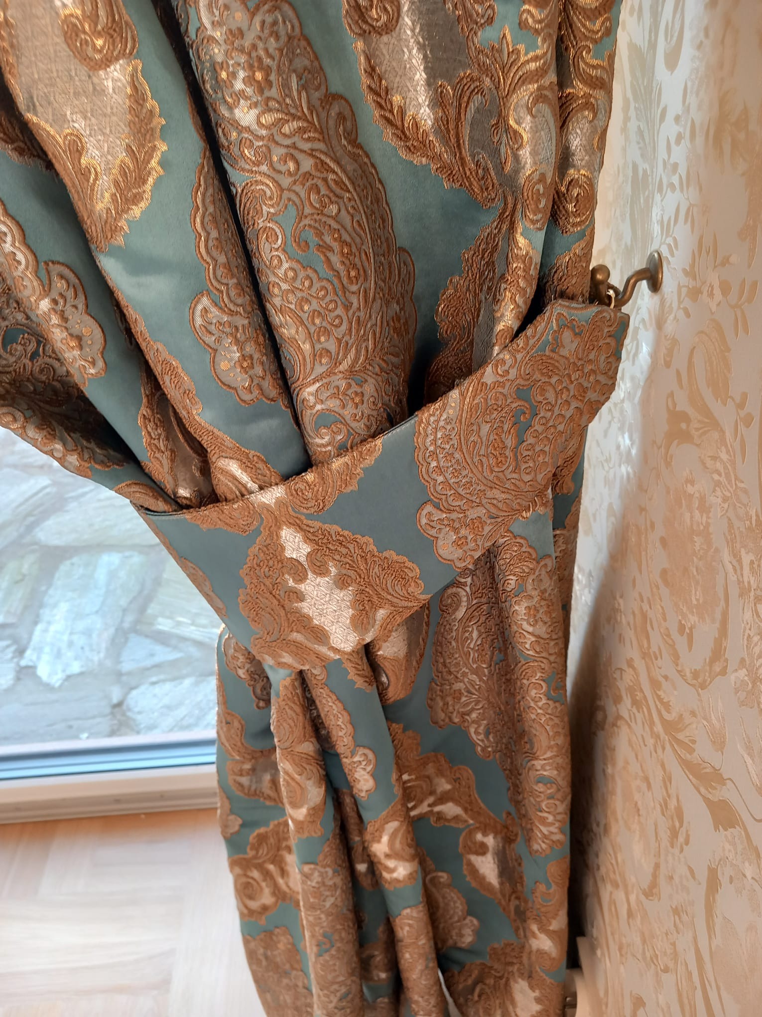 Turkusowa zasłona z ornamentem w kolorze złota, podpięta ozdobnym troczkiem, widoczna na tle ściany w kwiatowe wzory i fragment okna.