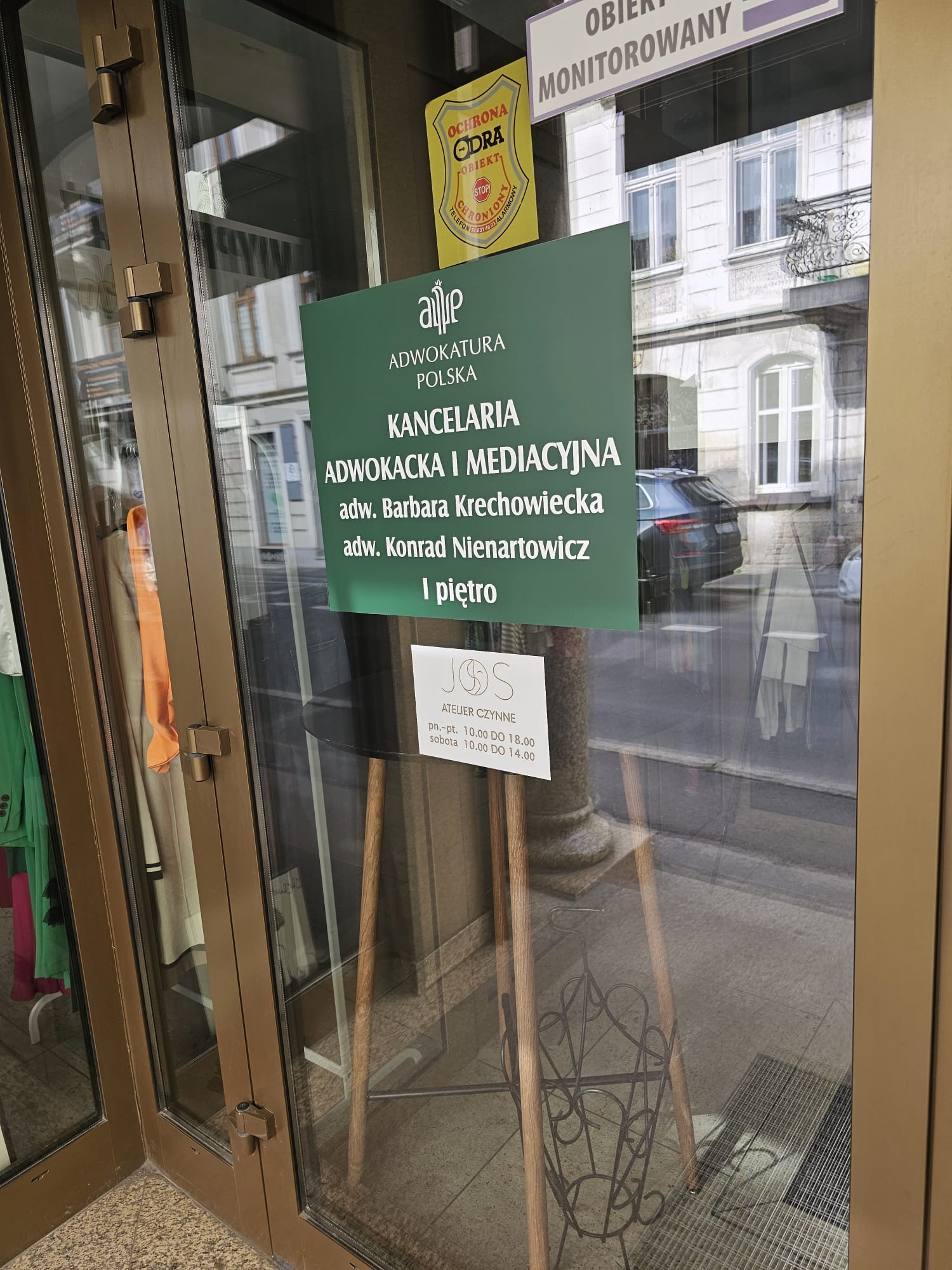 Znak informacyjny Kancelarii Adwokackiej i Mediacyjnej z nazwiskami adwokatów Barbara Krechowiecka i Konrad Nienartowicz, umieszczony na szklanych drzwiach wejściowych do budynku, widoczne odbicie...