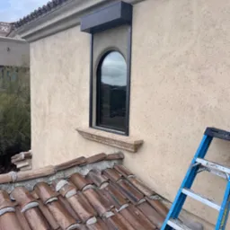 Zamknięta, czarna roleta nad owalnym oknem w beżowej ścianie z fakturą, widok z dachu pokrytego brązową dachówką, z boku oparta niebieska drabina.