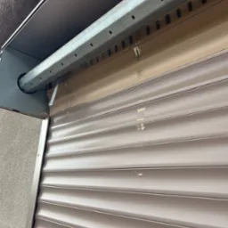 Częściowo zwinięta brązowa roleta zewnętrzna z widocznymi śladami użytkowania i zabrudzeniami na listwach.