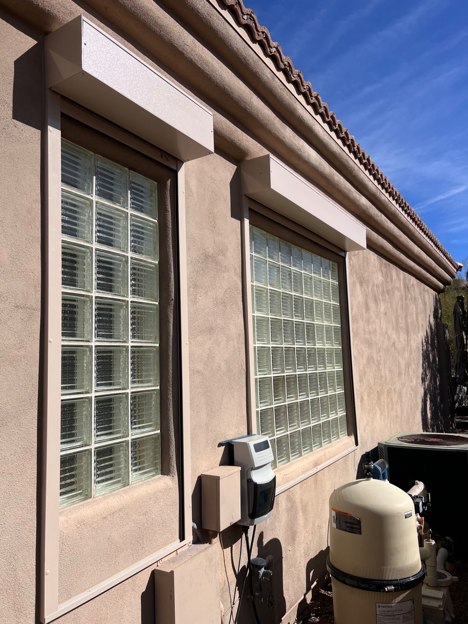 Zewnętrzne rolety z jasnego metalu zamontowane nad oknami z luksferami w budynku o teksturowanej, piaskowej elewacji i czerwonym dachu z dachówki ceramicznej.