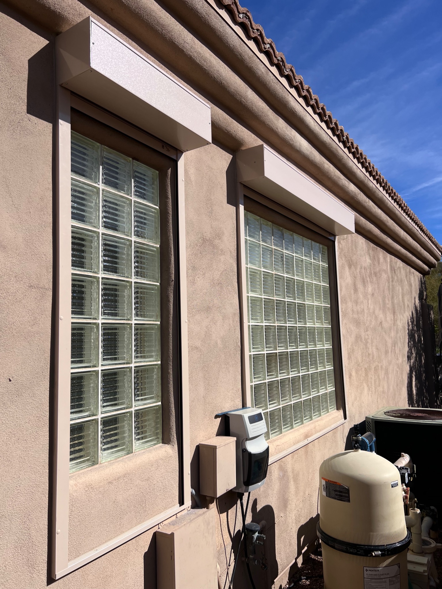 Zewnętrzne rolety w kasecie zamontowane nad oknami z luksferami na tle ściany budynku z terakotową dachówką