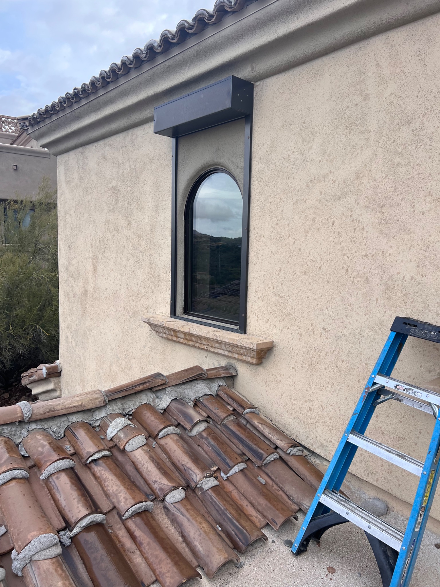 Zamknięta, czarna roleta nad owalnym oknem w beżowej ścianie z fakturą, widok z dachu pokrytego brązową dachówką, z boku oparta niebieska drabina.
