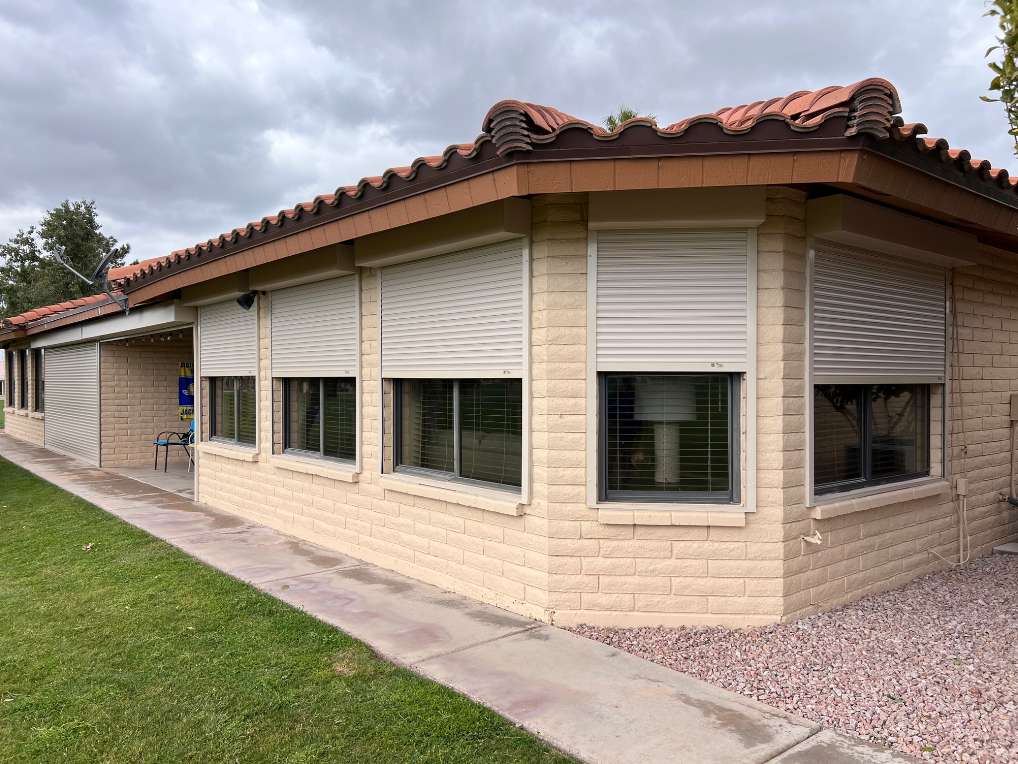 Beżowy dom z cegły z oknami zabezpieczonymi białymi roletami zwijanymi, brązowy dach z terakoty, pochmurne niebo.