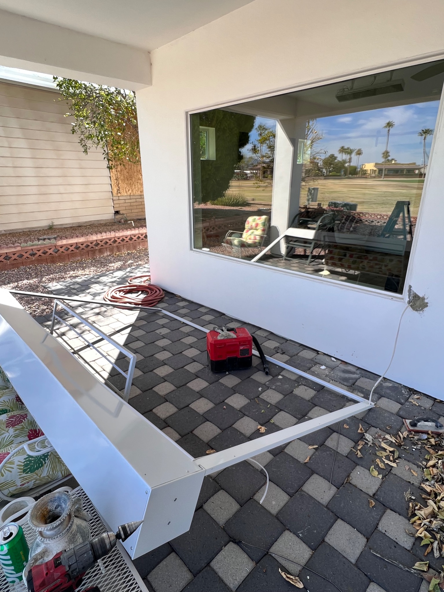Przygotowanie do montażu osłony okiennej: biała konstrukcja leży na kostce brukowej obok okna, widoczny zwinięty wąż ogrodowy, narzędzia i resztki po pracy.