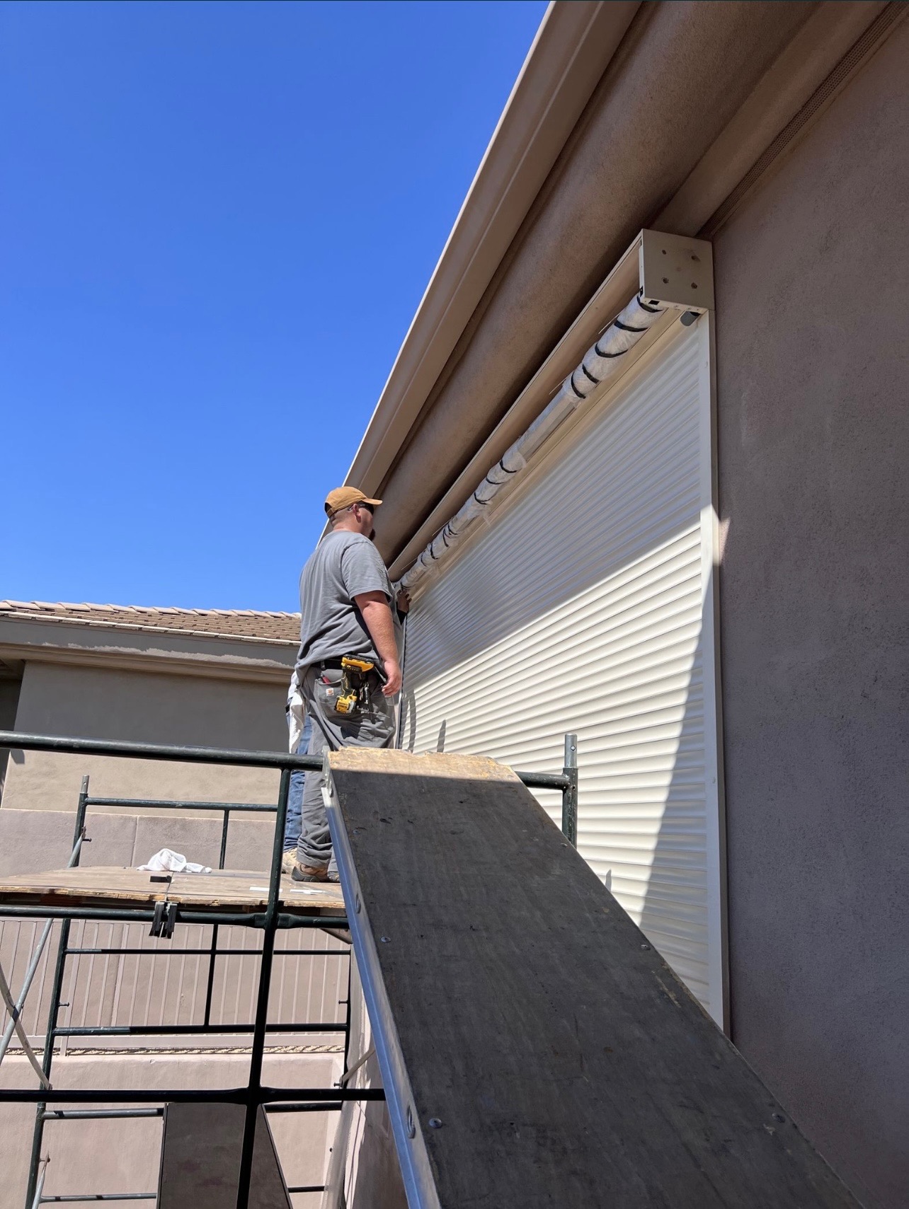 Montaż rolety zewnętrznej na elewacji domu przez pracownika stojącego na rusztowaniu, widoczne narzędzia w pasie.
