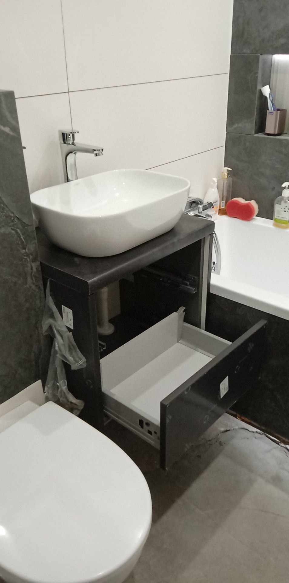 Umywalka nablatowa w łazience z otwartą szufladą szafki pod umywalką, widoczne białe płytki na ścianie i fragment toalety.