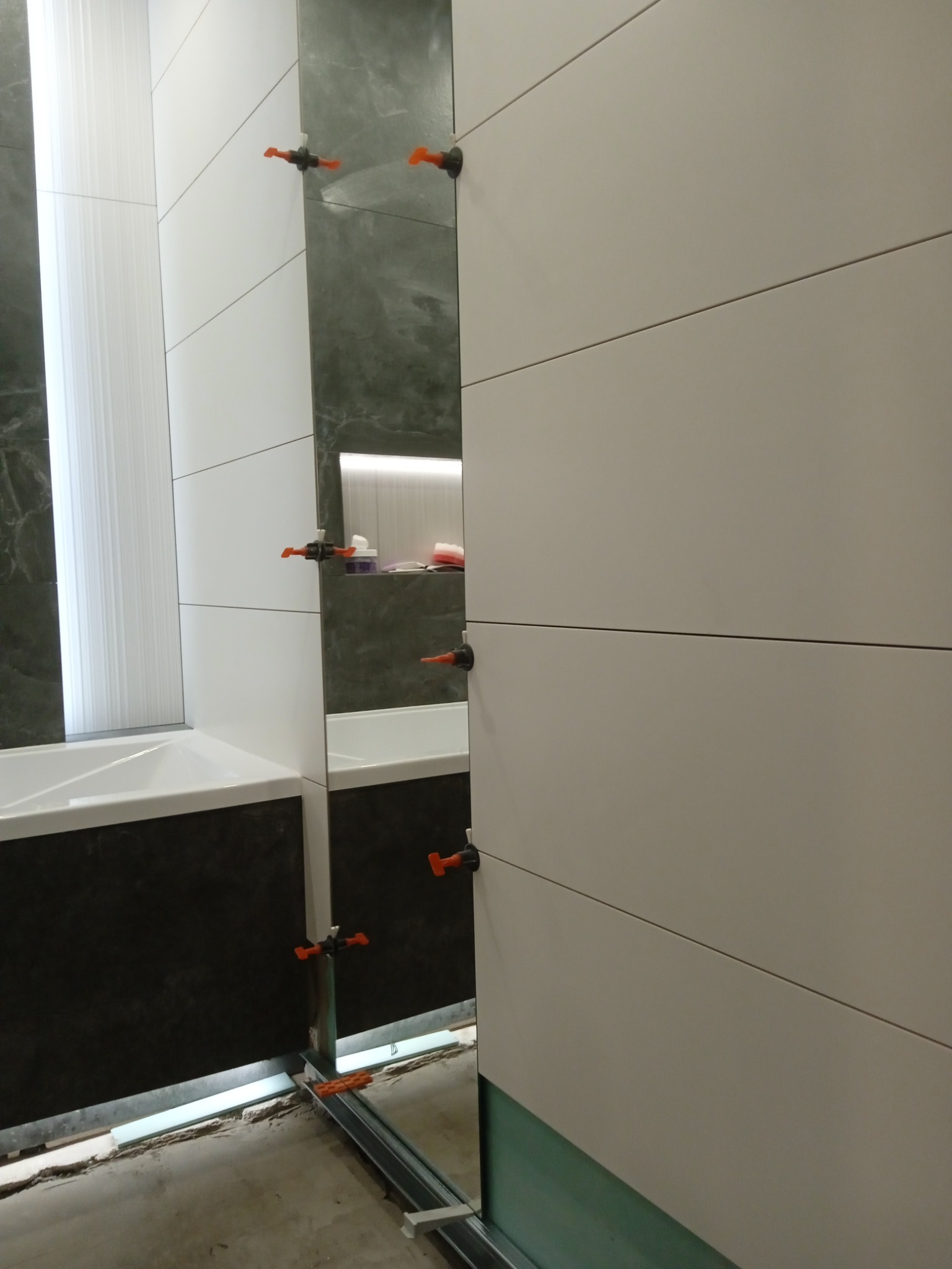 Układanie poziomych, białych płytek w łazience, widoczne pomarańczowe klipsy poziomujące, profil pod płytki i fragment wanny.