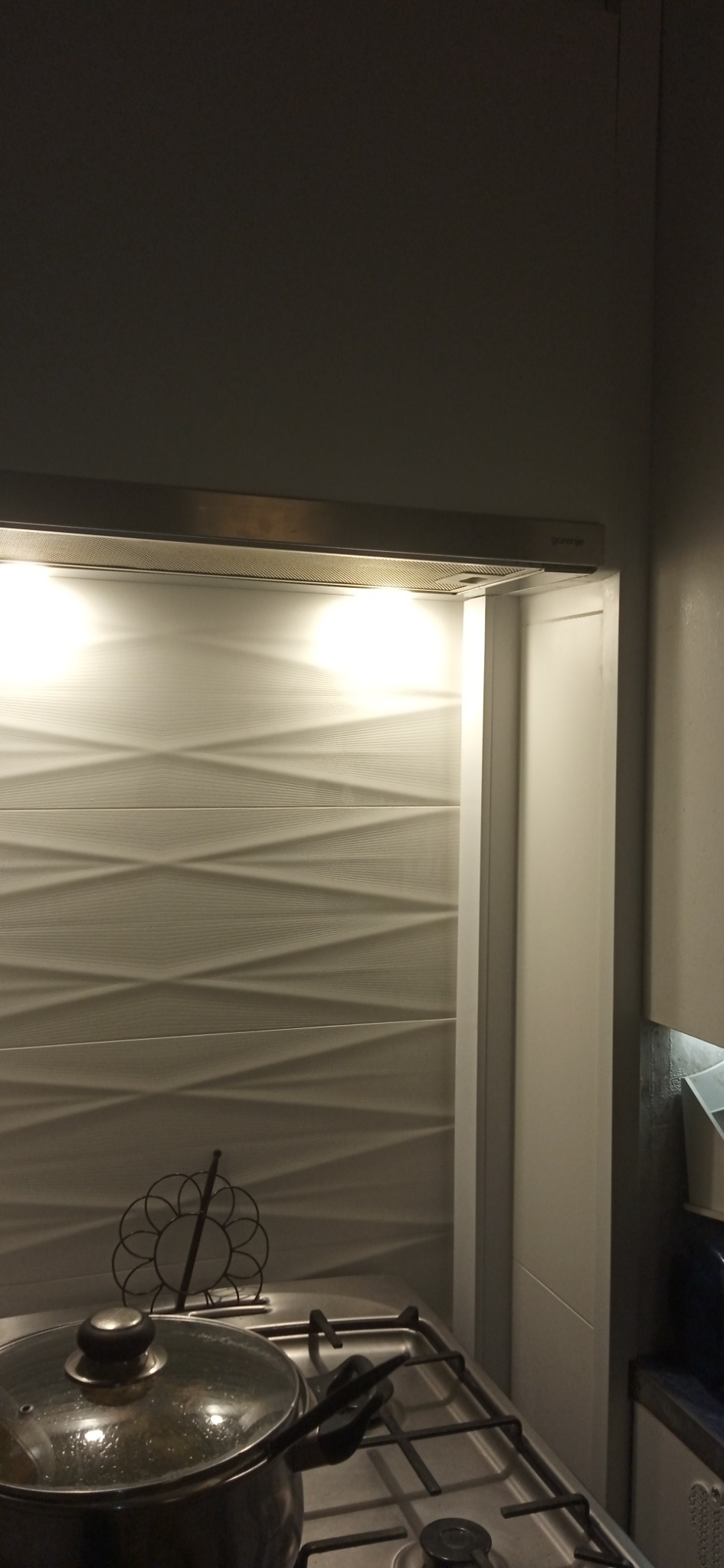 Kuchenny okap Gorenje z oświetleniem nad płytą gazową z garnkiem i ozdobnym stojakiem na ścianie z trójwymiarową fakturą.