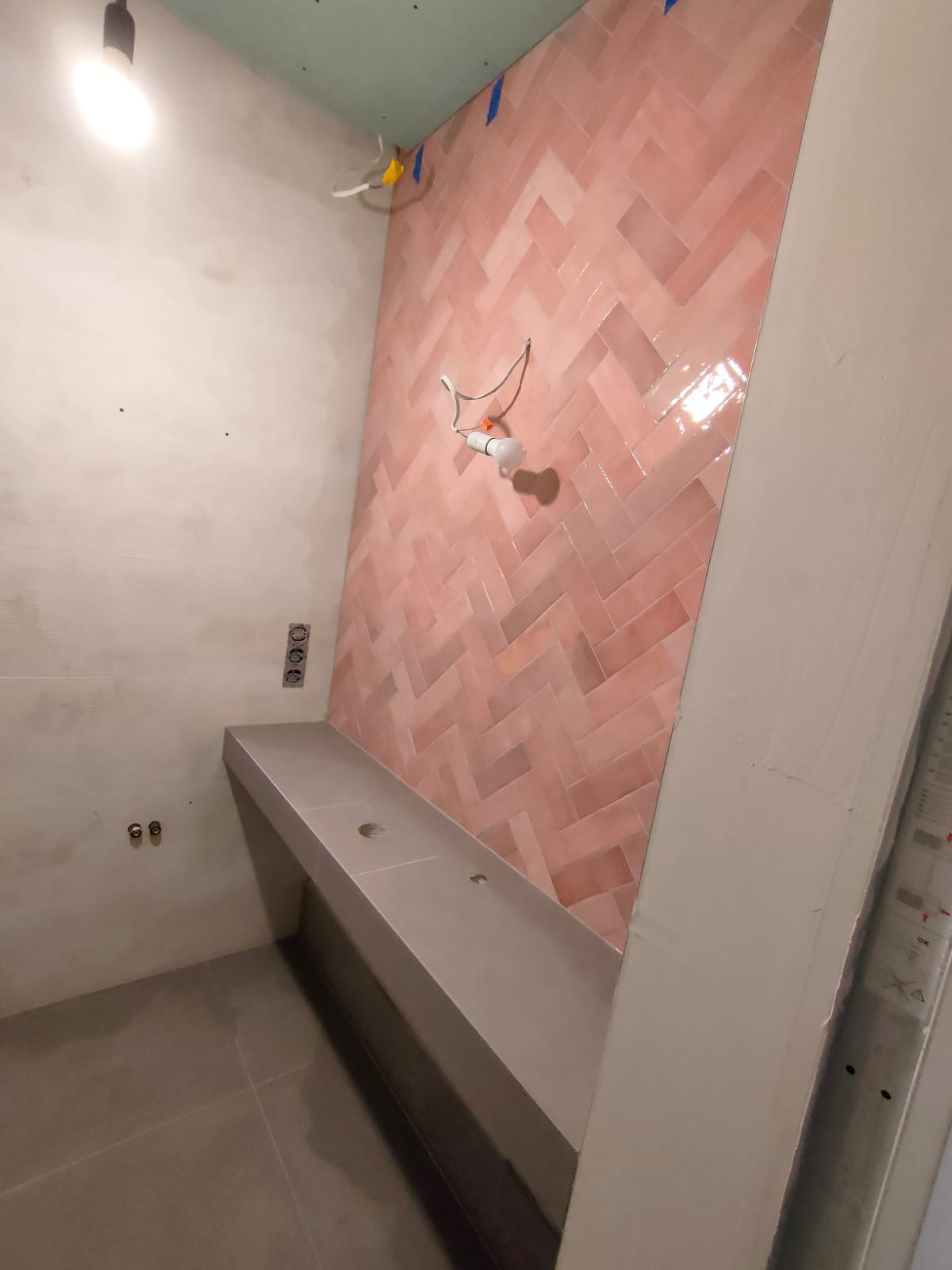 Ściana łazienki w Warszawie wyłożona błyszczącymi, różowymi płytkami ułożonymi w jodełkę, z widoczną instalacją elektryczną i częściowo zamontowaną umywalką podwieszaną.