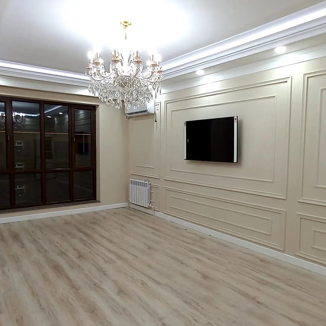 Jasne, puste pomieszczenie z panelami ściennymi, drewnianą podłogą, kryształowym żyrandolem i telewizorem na ścianie.