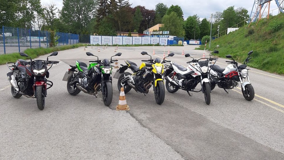 Rząd motocykli różnych marek i kolorów zaparkowanych na placu manewrowym z pomarańczowymi pachołkami, teren treningowy.