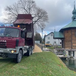 Sprzedaż Materiałów Stanisław Wojciechowski - Ciężarówka Tatra z podniesioną skrzynią wyładowuje piasek na trawniku obok drewnianego budynku z zielonym dachem, w tle widoczna koparka.